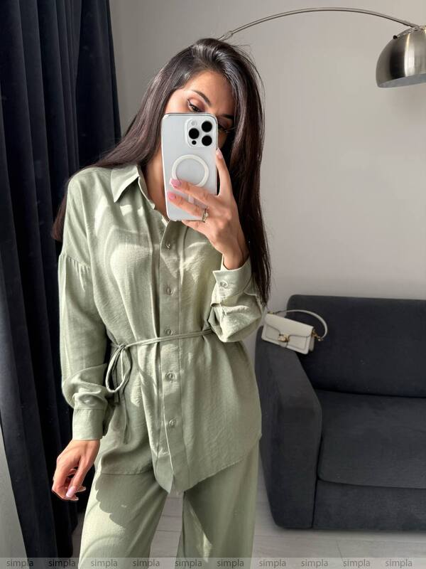 Woman taking a mirror selfie wearing a green outfit in a room with a gray sofa and lamp.