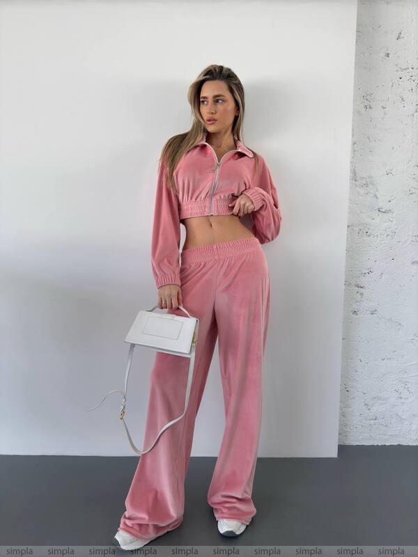 Woman wearing a pink tracksuit holding a white handbag against a white wall.