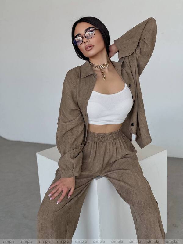 Woman wearing a brown outfit with a white top, sitting on a white cube against a plain background.