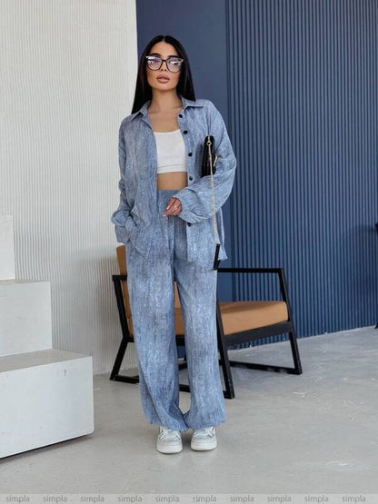 Woman wearing a blue denim outfit standing in an indoor setting with a chair and wall in the background.