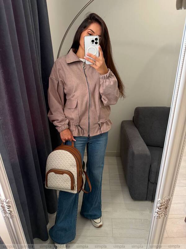 Person wearing a beige jacket and blue jeans, holding a small backpack, standing in front of a mirror.
