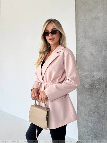 Woman wearing a pink coat and sunglasses, holding a beige handbag against a white and gray wall.