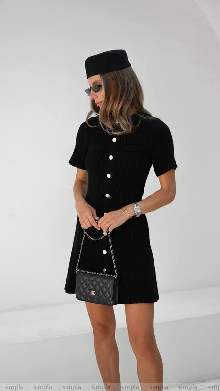 Woman wearing a black dress with white buttons and a matching black hat, holding a black handbag.
