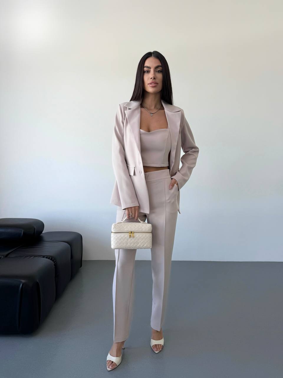 Woman in a beige suit with a handbag standing in a minimalistic room.