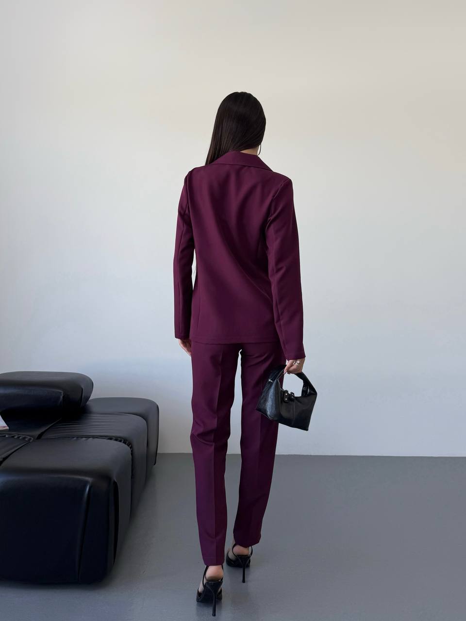 Person wearing a burgundy suit holding a black handbag in a minimalistic room.