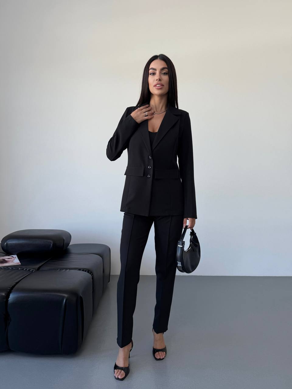 Woman in a black suit standing in a minimalistic room with a black sofa.