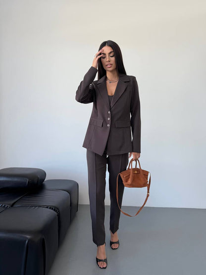 Woman in a dark suit holding a brown handbag against a plain background