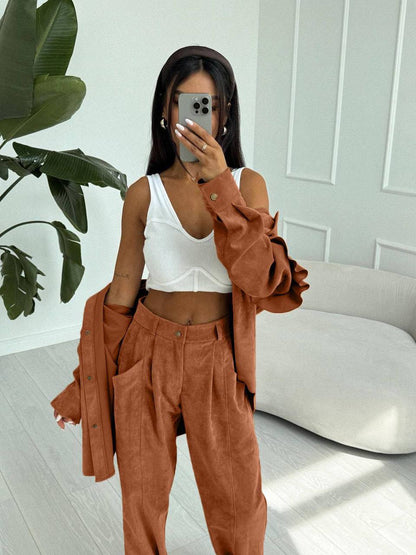 Person wearing a white top and brown pants with a jacket in a room with a plant and white wall.