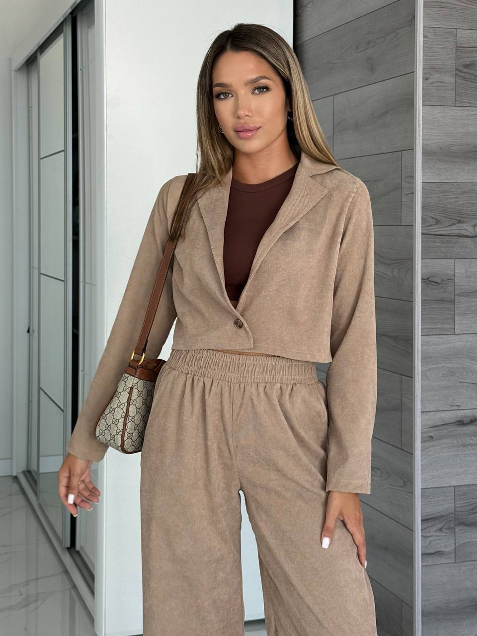 Woman wearing a beige outfit with a brown top, standing against a neutral wall.