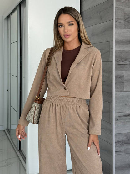 Woman wearing a beige outfit with a brown top, standing against a neutral wall.