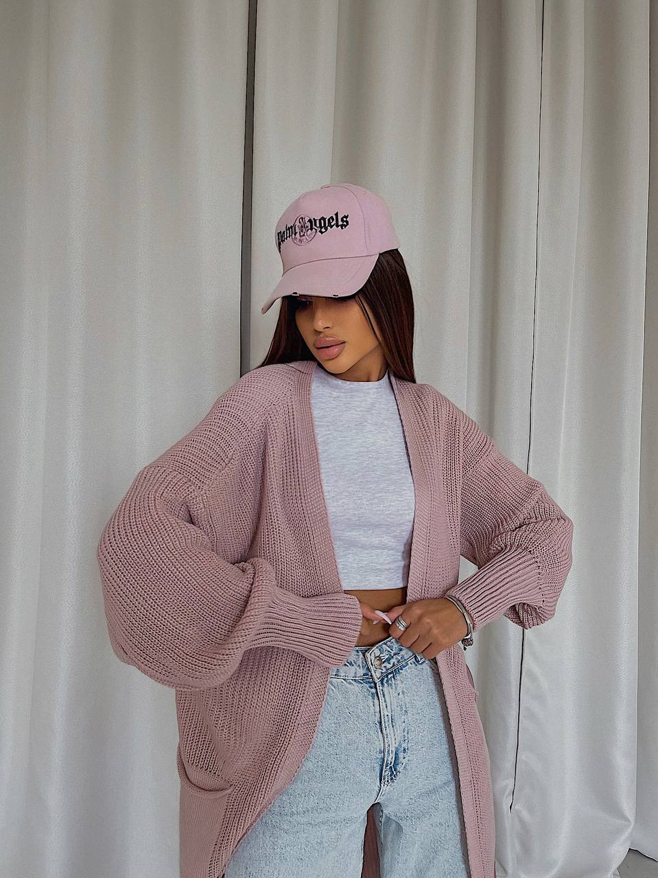 Person wearing a pink cardigan, light blue jeans, and a pink cap with text against a white curtain background