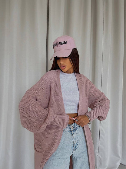Person wearing a pink cardigan, light blue jeans, and a pink cap with text against a white curtain background