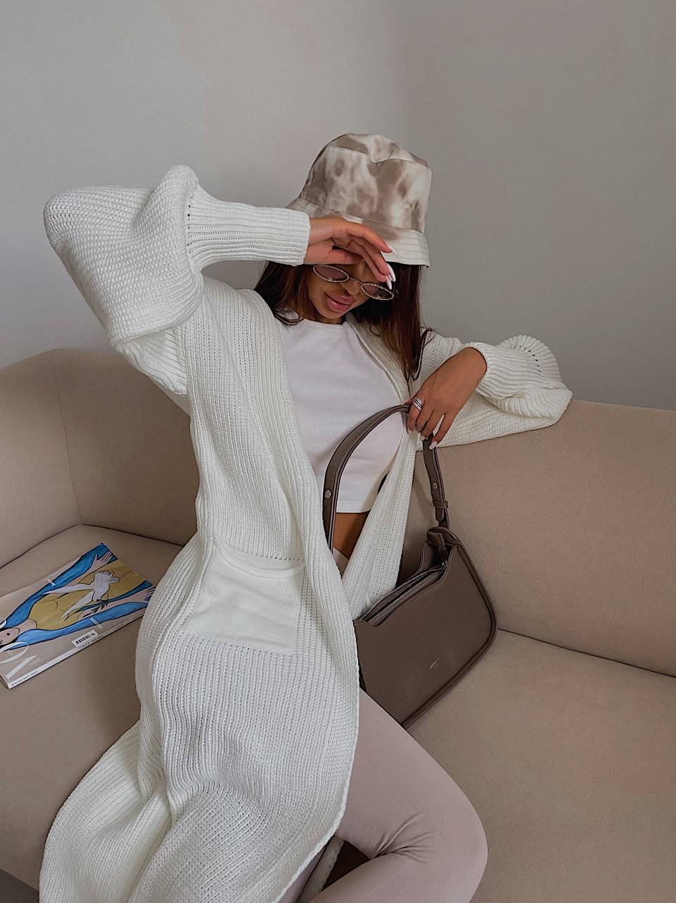 Person sitting on a couch wearing a white sweater and hat, holding a brown handbag.