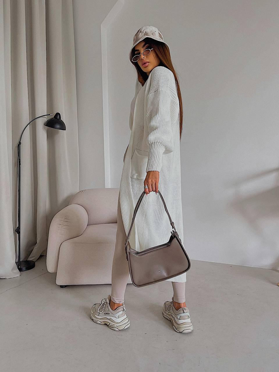 Woman holding a gray handbag in a minimalistic room with a beige armchair and lamp.