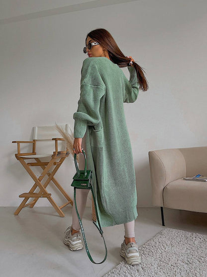 Woman in a long green cardigan holding a green handbag in a room with a chair and sofa.