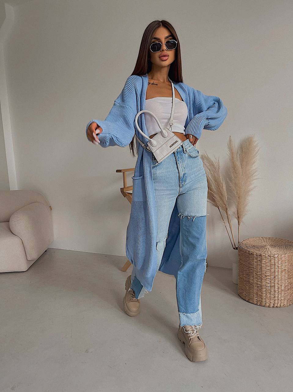 Woman in a stylish outfit with a blue cardigan and jeans, holding a white handbag, in a minimalistic room.