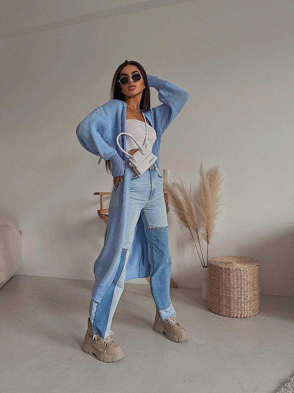 Woman wearing a blue cardigan, white top, and jeans in a minimalistic room.