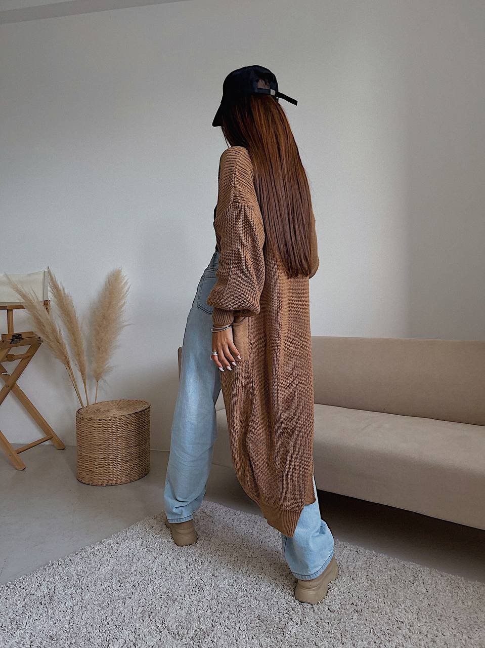 Person wearing a long brown cardigan and light blue jeans standing in a room with a beige sofa and wicker basket.