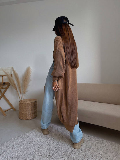 Person wearing a long brown cardigan and light blue jeans standing in a room with a beige sofa and wicker basket.