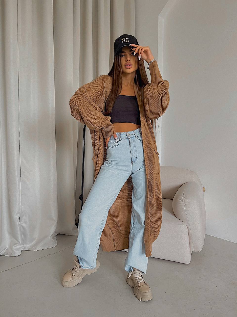 Woman wearing a brown cardigan, black top, light blue jeans, and beige sneakers in a minimalistic room.