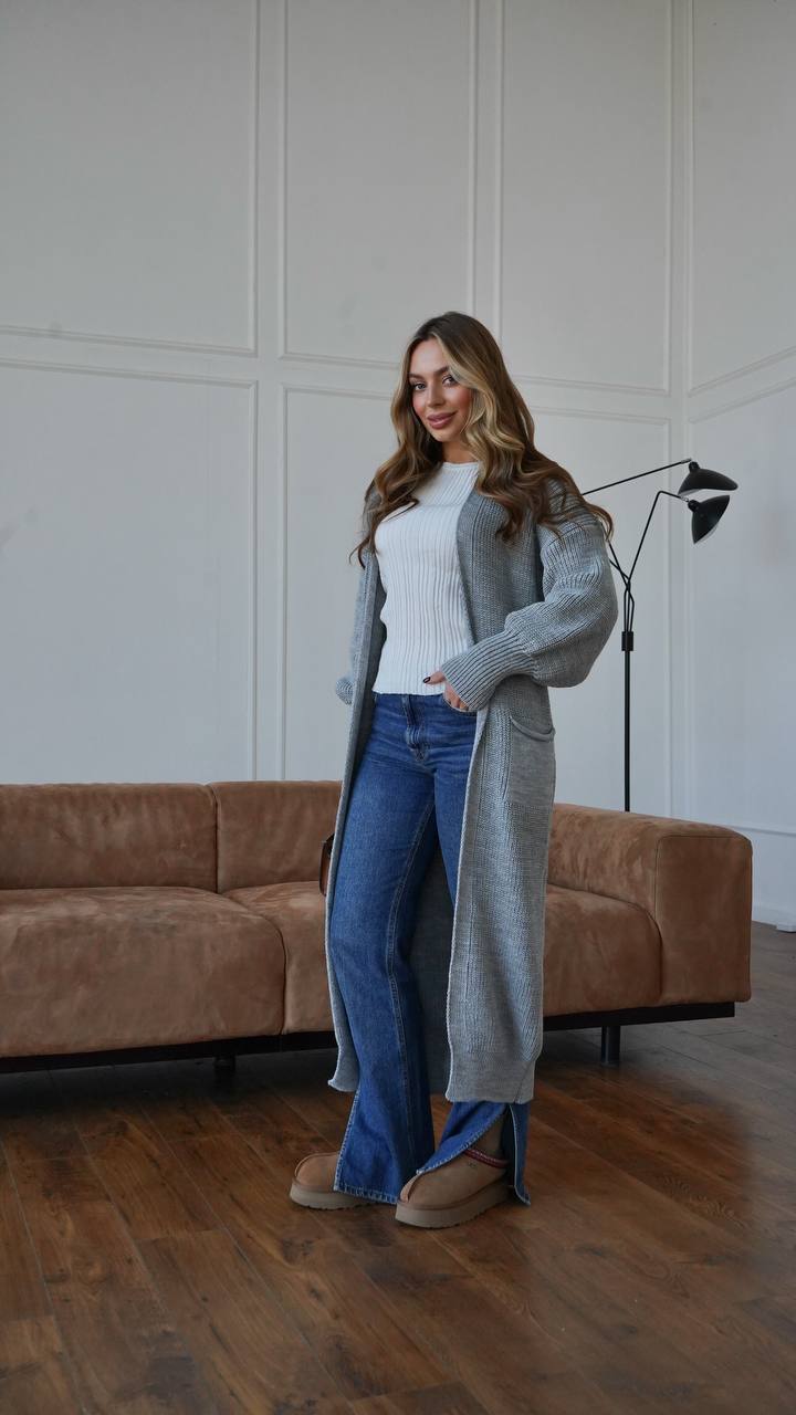 Woman in a gray cardigan and blue jeans standing in a room with a brown sofa and white walls.