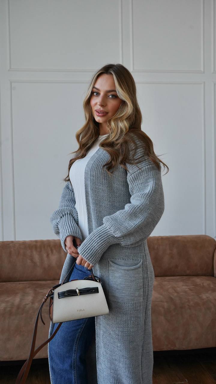 Woman in a long gray cardigan holding a white handbag against a neutral background