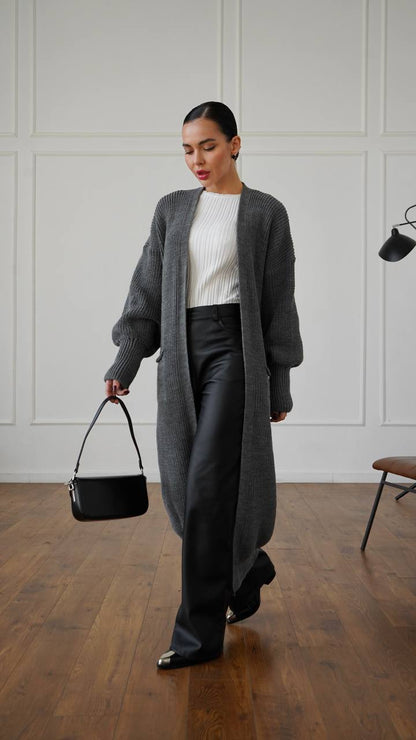 Woman in a gray cardigan and black pants holding a black handbag in a minimalistic room.