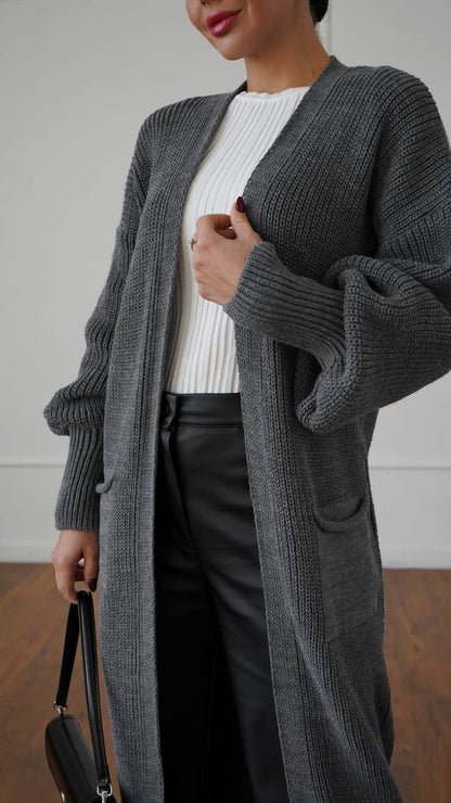 Person wearing a long gray cardigan over a white sweater and black pants, holding a black handbag.