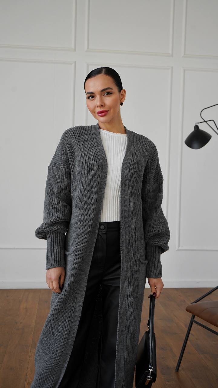 Woman wearing a long gray cardigan over a white top, standing indoors against a white paneled wall.