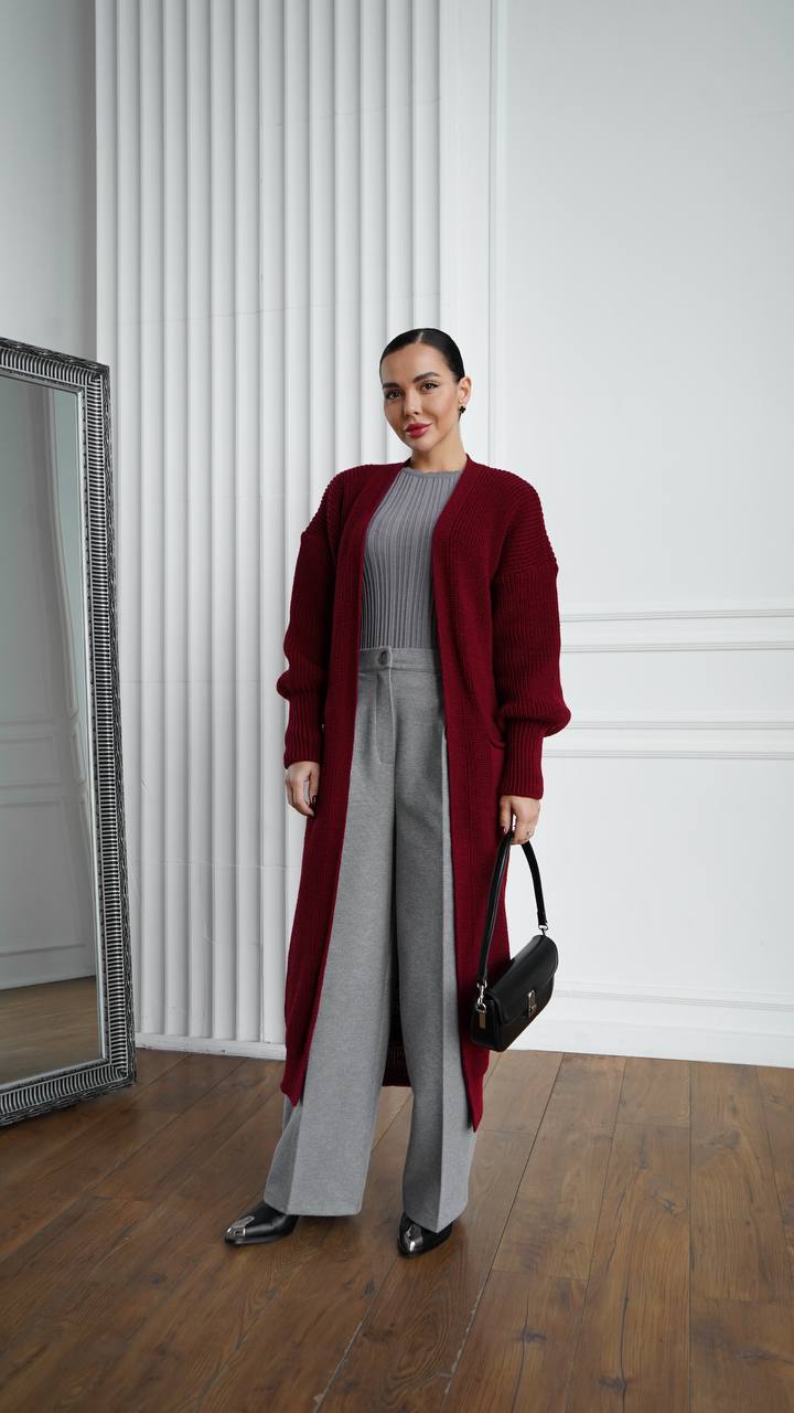 Woman wearing a red cardigan and gray pants standing in a room with a mirror and white walls.