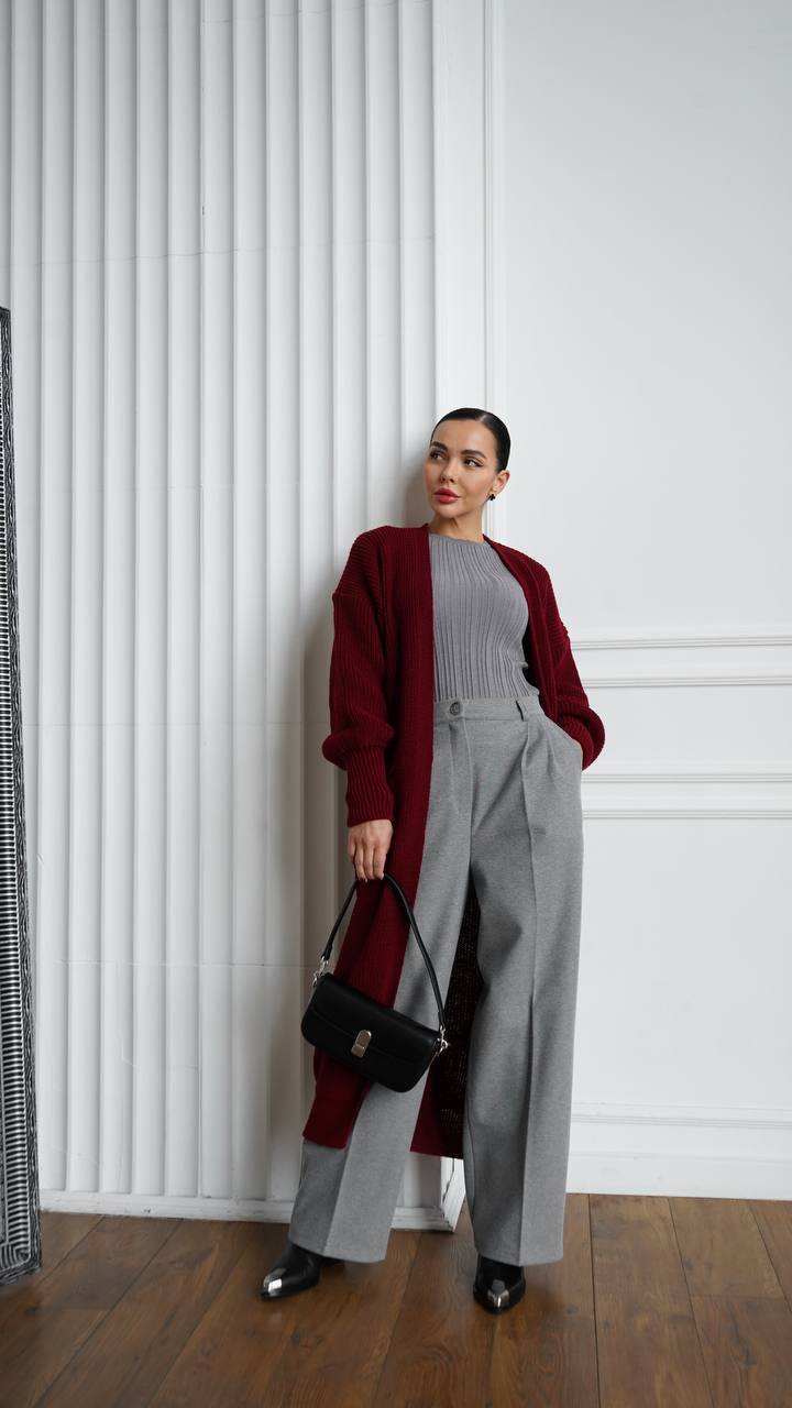 Woman wearing a red cardigan, gray sweater, and wide-leg pants against a white wall.