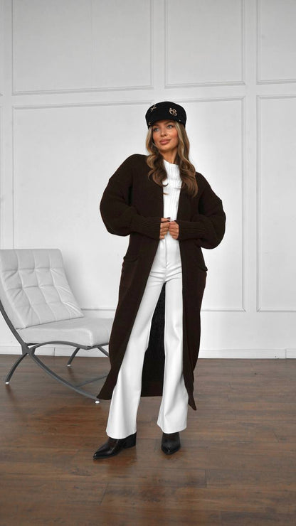 Woman wearing a brown long coat and white pants standing in a room with a white wall and gray chair.