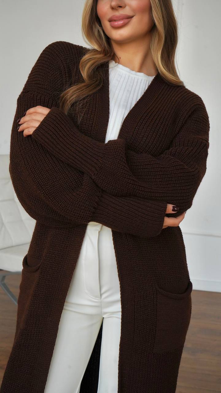 Woman wearing a brown cardigan over a white top and pants, standing against a neutral background.
