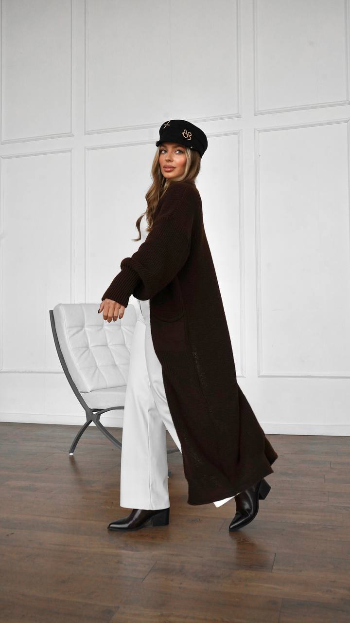 Woman wearing a long brown coat and white pants in a minimalistic room.