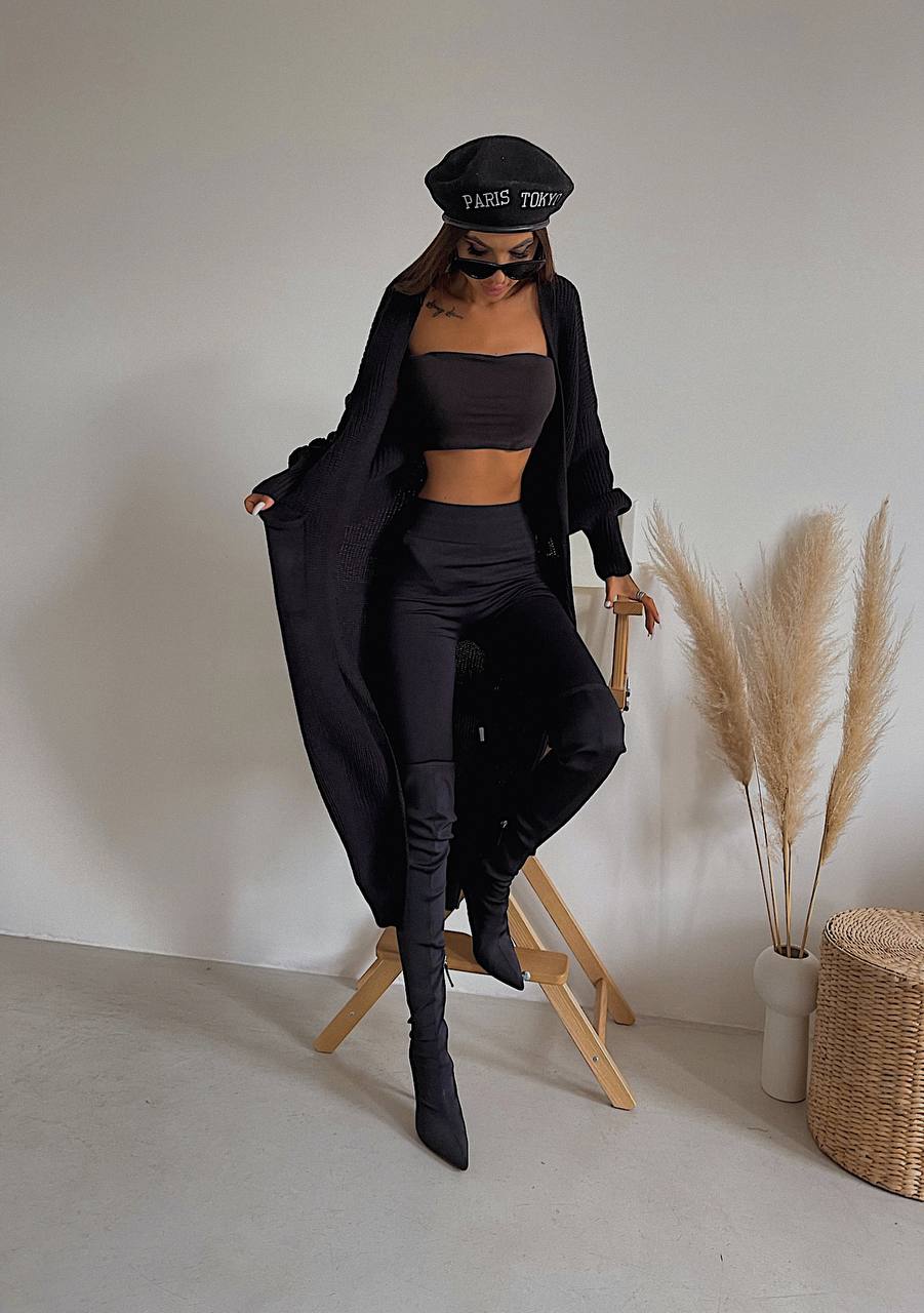 Person wearing a black outfit with a hat and sunglasses, standing on a wooden stool against a white wall.
