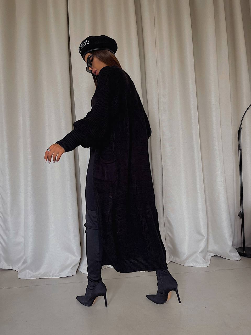 Person wearing a long black coat and black shoes in front of white curtains