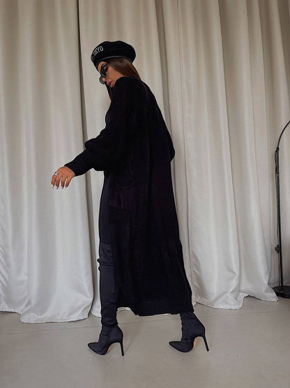 Person wearing a long black coat and black shoes in front of white curtains