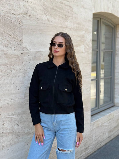 Woman wearing a black jacket and light blue jeans standing against a beige wall.