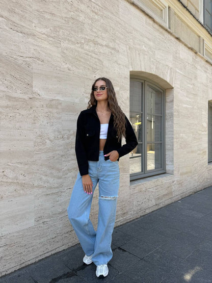 Woman in black jacket, white top, and light blue jeans standing against a stone wall.