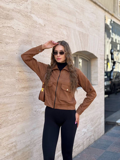 Woman in a brown jacket and black leggings standing against a stone wall.