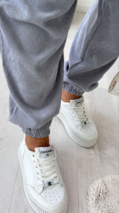 White sneakers with a brand logo worn with gray pants on a light wooden floor.