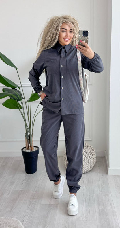Person taking a mirror selfie wearing a dark gray outfit with white sneakers in a room with a plant and rug.