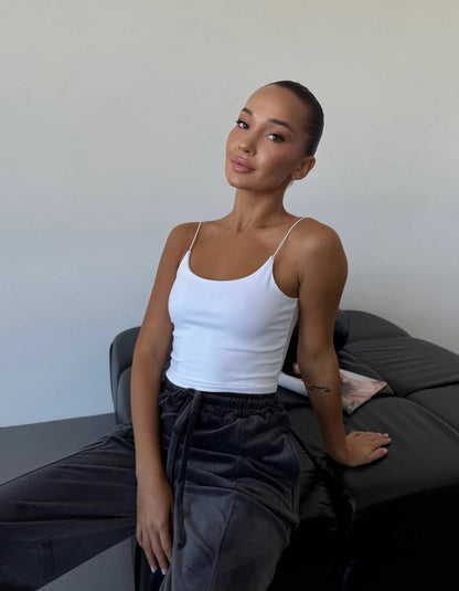 Woman sitting on a bed wearing a white tank top and dark pants against a plain background