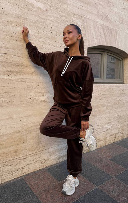Person wearing a brown tracksuit leaning against a stone wall.