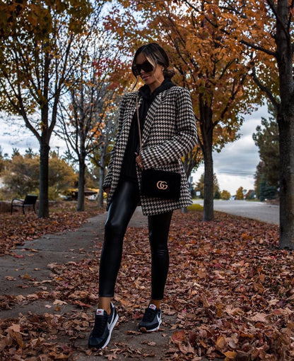 Person wearing a houndstooth coat and black leggings standing on a leaf-covered path with trees in the background.