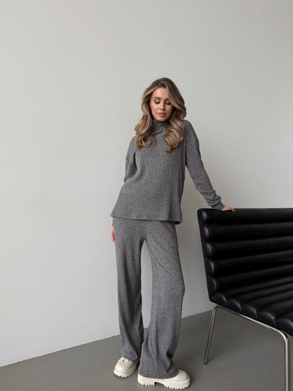 Woman wearing a gray outfit standing next to a black chair against a plain wall.