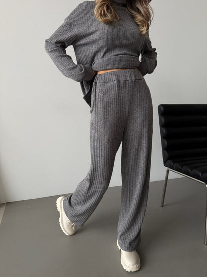 Person wearing a gray knit outfit standing in a minimalistic room with a chair.