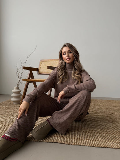 Woman sitting on a rug wearing a brown sweater and pants in a minimalistic room.