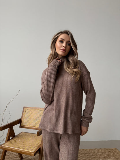 Woman wearing a brown ribbed sweater and pants standing in a room with a chair and decorative branch.