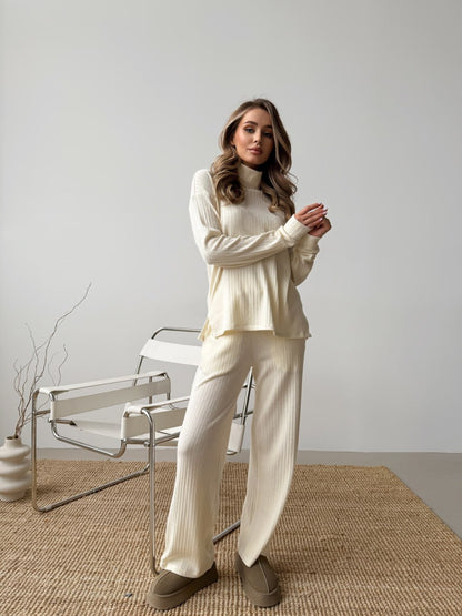 Woman wearing a cream-colored outfit standing in a minimalistic room with a chair and vase.